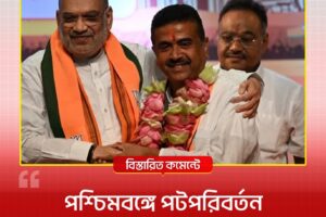 Two men on stage smiling and embracing; the man on the left wears an orange scarf, the man in the center has a pink flower garland around his neck, with a third person in the background